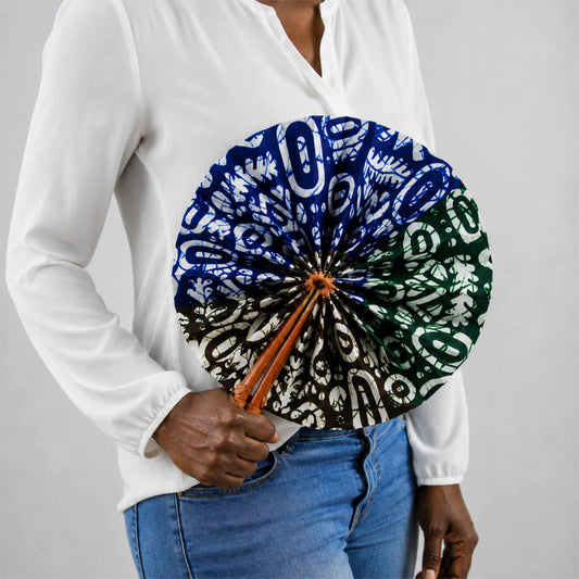 Éventail africain artisanal rond en wax bleu, vert, marron et blanc, présenté ouvert et tenu à la main avec une blouse blanche et un jean.