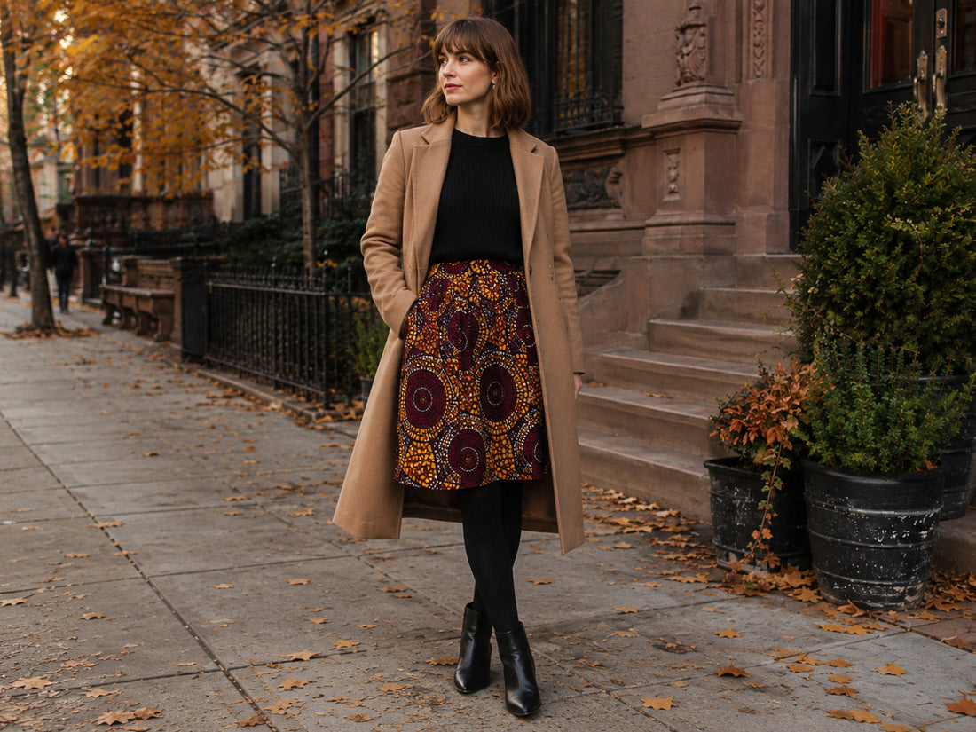 Femme portant une jupe wax avec pull noir, manteau camel et bottines pour un look automne-hiver