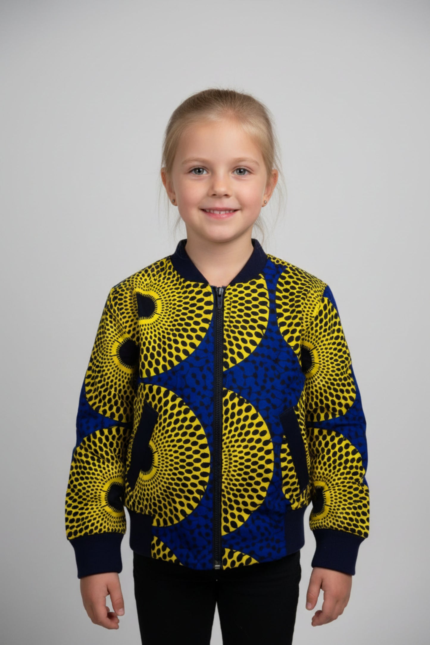 Fillette souriante portant un bomber jacket en tissu wax jaune citron à grands motifs cercles concentriques noirs et intercalaires bleu roi avec col et poignets côtelés bleu marine - Vêtement enfant africain Djêkoo Wax