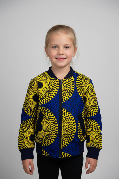 Fillette souriante portant un bomber jacket en tissu wax jaune citron à grands motifs cercles concentriques noirs et intercalaires bleu roi avec col et poignets côtelés bleu marine - Vêtement enfant africain Djêkoo Wax