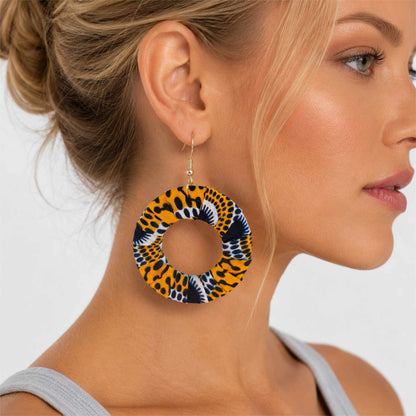 Boucles d'oreilles pendantes artisanales en tissu wax africain, forme cercle ajouré, motif léopard jaune soleil à pois noirs et arabesques marine — portées en situation