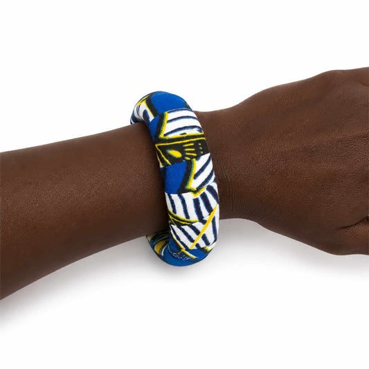 Bracelet africain artisanal large recouvert de tissu wax bleu, blanc, noir et jaune, présenté porté au poignet sur fond blanc.