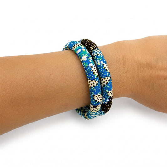 Duo de bracelets joncs rigides recouverts de tissu wax bleu aux motifs géométriques et pois noirs, porté au poignet sur fond blanc.
