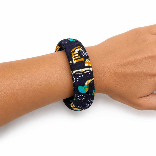 Bracelet jonc large artisanal recouvert de tissu wax bleu marine, blanc, jaune et turquoise, présenté porté au poignet sur fond blanc.