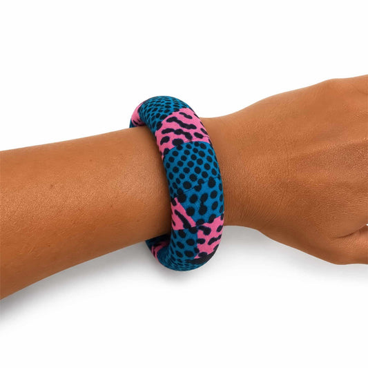 Bracelet jonc large artisanal recouvert de tissu wax bleu turquoise, rose et noir à motifs graphiques, présenté porté au poignet sur fond blanc.