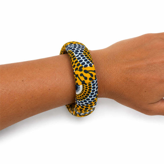 Bracelet jonc large artisanal recouvert de tissu wax jaune, noir et blanc à motifs graphiques et pois, présenté porté au poignet sur fond blanc.