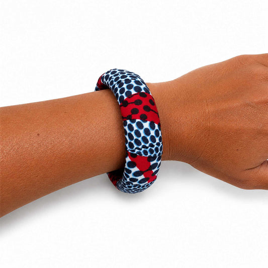 Bracelet jonc large artisanal recouvert de tissu wax rouge, blanc et bleu marine à motifs de pois, présenté porté au poignet sur fond blanc.