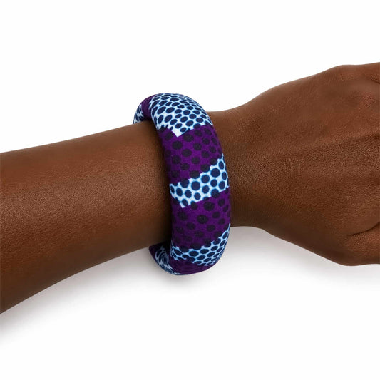 Bracelet jonc large artisanal recouvert de tissu wax violet, blanc et bleu marine à motifs de pois, présenté porté au poignet sur fond blanc.