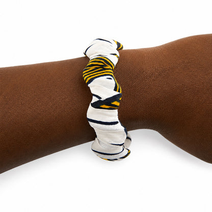 Bracelet jonc ondulé en bois recouvert de tissu wax blanc à rayures noires et jaunes, porté au poignet sur fond blanc.