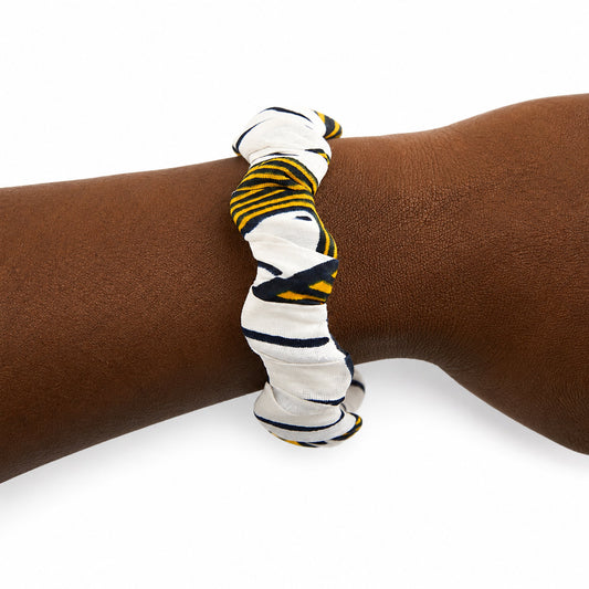 Bracelet jonc ondulé en bois recouvert de tissu wax blanc à rayures noires et jaunes, porté au poignet sur fond blanc.