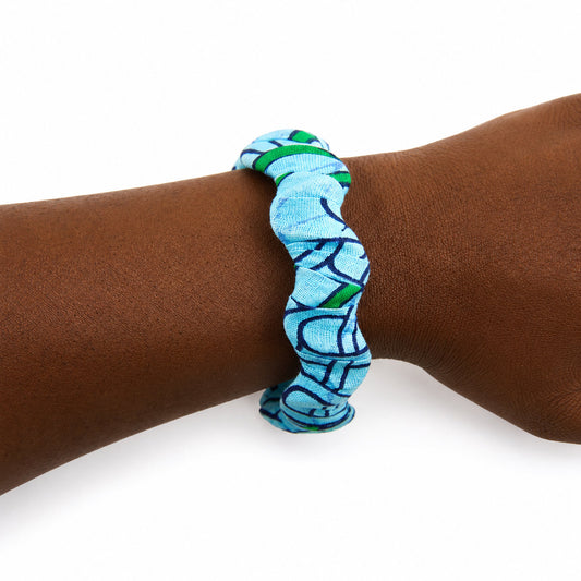 Bracelet jonc ondulé en bois recouvert de tissu wax bleu ciel aux motifs géométriques bleu marine et touches de vert, porté au poignet sur fond blanc.