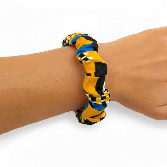 Bracelet jonc ondulé en bois recouvert de tissu wax jaune vibrant aux motifs géométriques complexes (damiers noirs, bandes bleu royal), porté au poignet sur fond blanc.