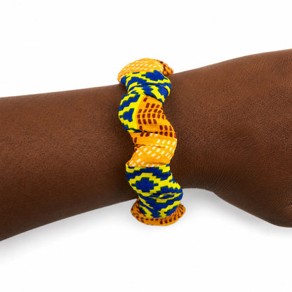 Bracelet jonc ondulé en bois recouvert de tissu wax aux motifs géométriques jaune-vert/bleu et texture orange/marron, porté au poignet sur fond blanc.