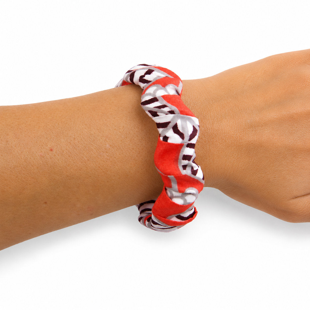 Bracelet jonc ondulé en bois recouvert de tissu wax rouge, blanc et gris aux motifs graphiques noirs, porté au poignet sur fond blanc.