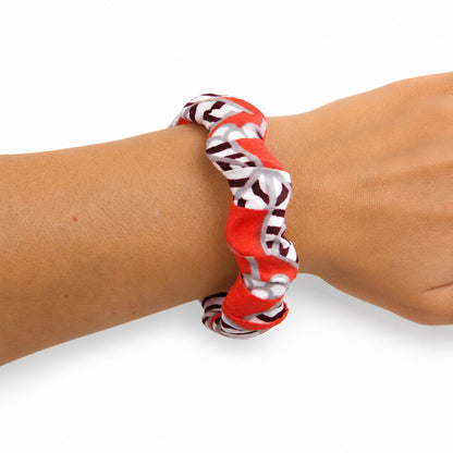 Bracelet jonc ondulé en bois recouvert de tissu wax rouge, blanc et gris aux motifs graphiques noirs, porté au poignet sur fond blanc.