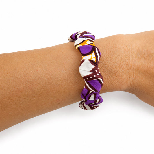 Bracelet jonc ondulé en bois recouvert de tissu wax violet et orange aux motifs graphiques, porté au poignet sur fond blanc.