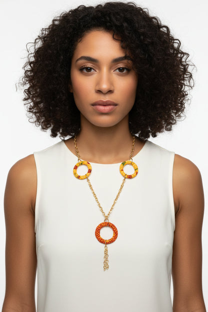 Femme portant un collier sautoir doré avec anneaux recouverts de tissu wax jaune orangée et grand anneau central orange rouge avec chaîne pendante - Bijou artisanal africain Djêkoo Wax