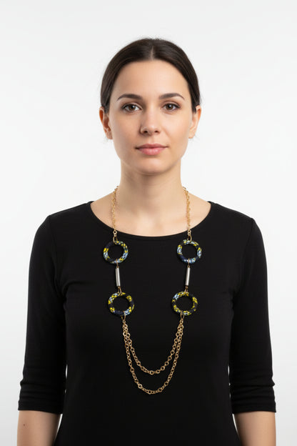 Femme portant un collier sautoir doré avec anneaux recouverts de tissu wax noir à motifs jaune citron et bleu turquoise, embouts cylindriques dorés et double chaîne pendante - Bijou artisanal africain Djêkoo Wax