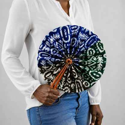 Éventail africain artisanal rond en wax bleu, vert, marron et blanc, présenté ouvert et tenu à la main avec une blouse blanche et un jean.