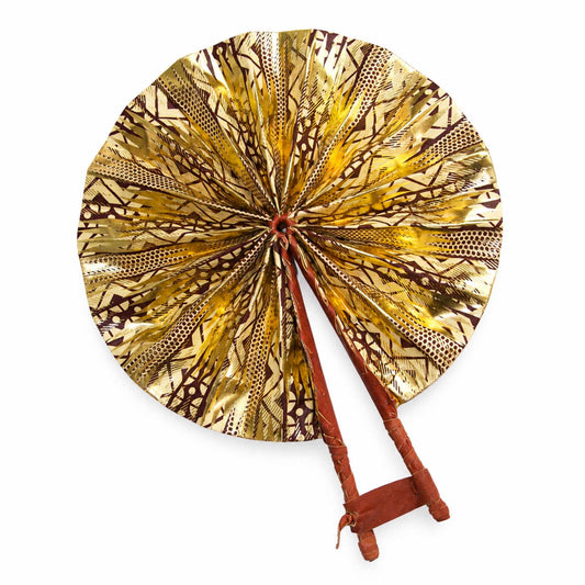 Éventail africain artisanal ouvert en tissu wax doré, beige et marron, avec motifs graphiques et structure en bois, sur fond blanc.