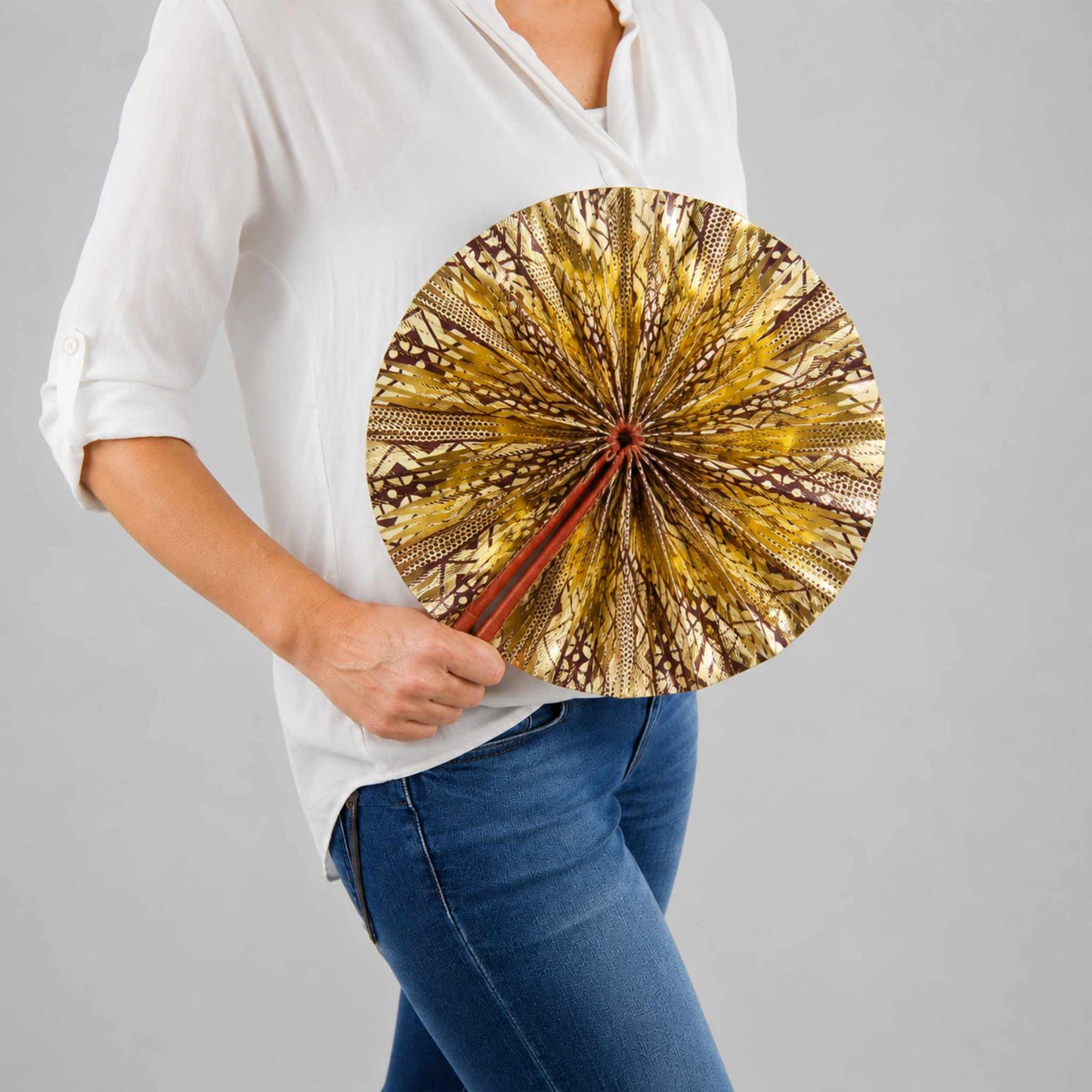 Éventail africain artisanal rond en wax doré, beige et marron, présenté ouvert et tenu à la main avec une blouse blanche et un jean.