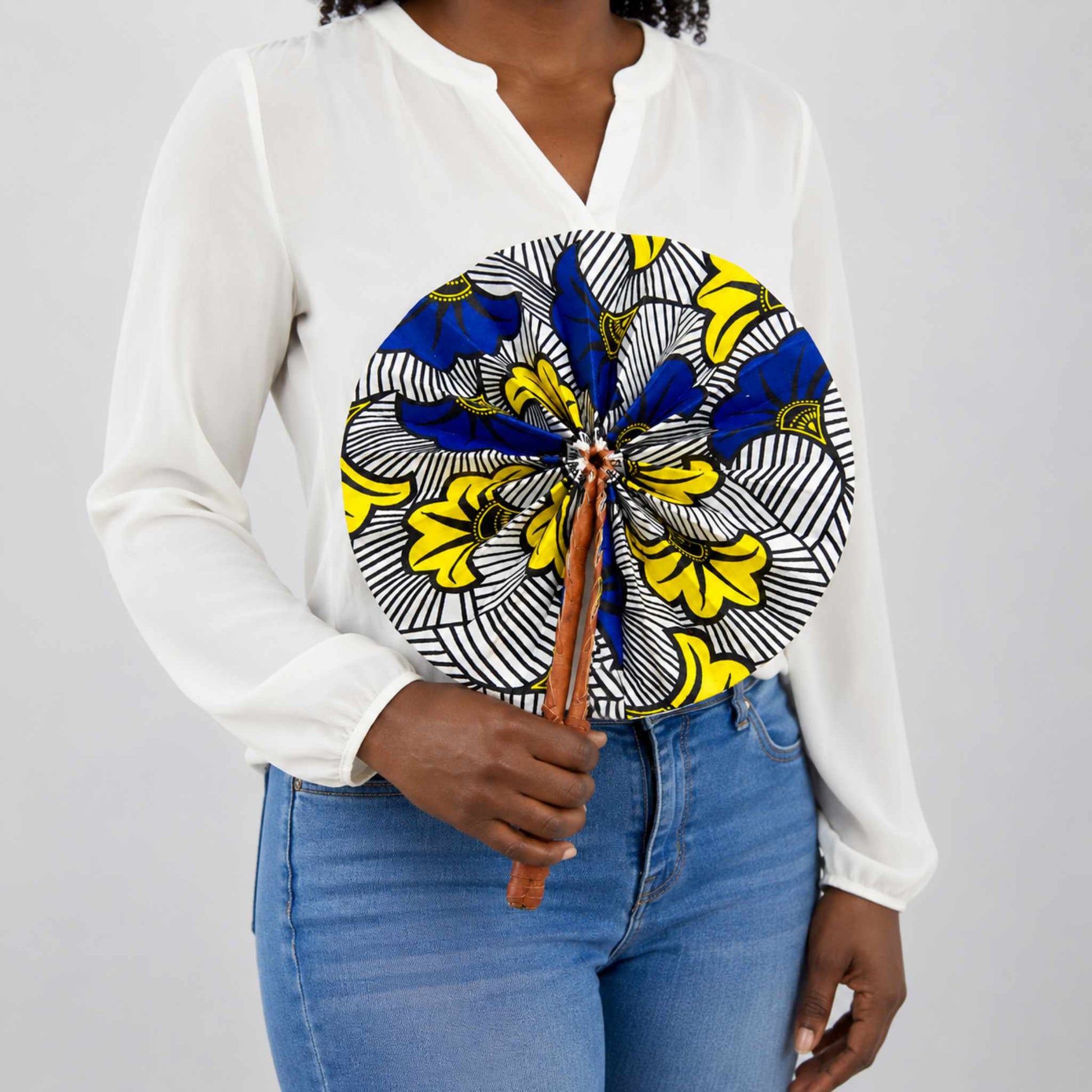 Éventail africain artisanal rond en wax bleu, jaune, noir et blanc, motif Fleurs de mariage, tenu à la main avec une blouse blanche.