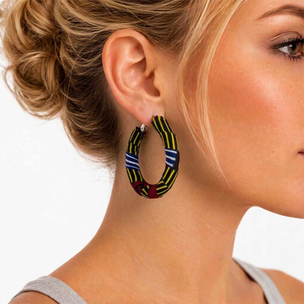Gros plan de profil d'une femme portant une boucle d'oreille créole recouverte de tissu wax à rayures jaunes, bleues et rouges.