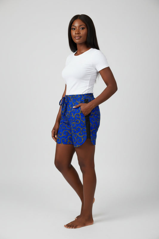 Femme portant un short en tissu wax bleu royal à motifs arabesques jaune doré pointillés avec ceinture élastique nœud bleu, bande latérale noire et poches latérales - Mode africaine femme Djêkoo Wax